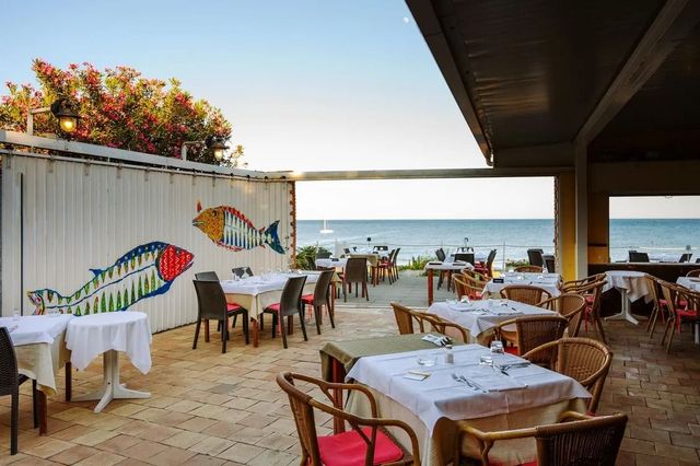 Terrazza del caffè sul mare con tavoli, sedie e un murale raffigurante un pesce dipinto, con vista sull'oceano al tramonto.