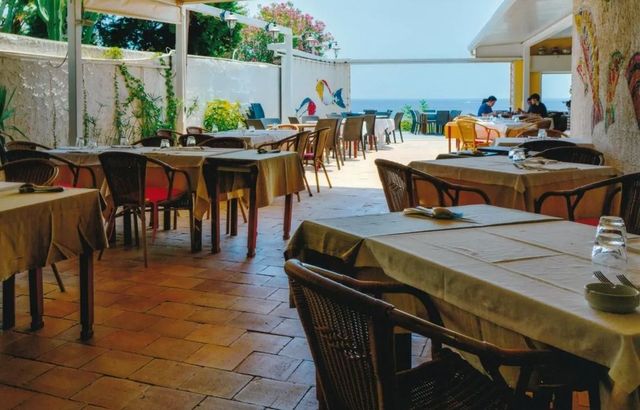 Ristorante all'aperto sul mare con tavoli vuoti, sedie in vimini e una vista soleggiata sull'oceano.