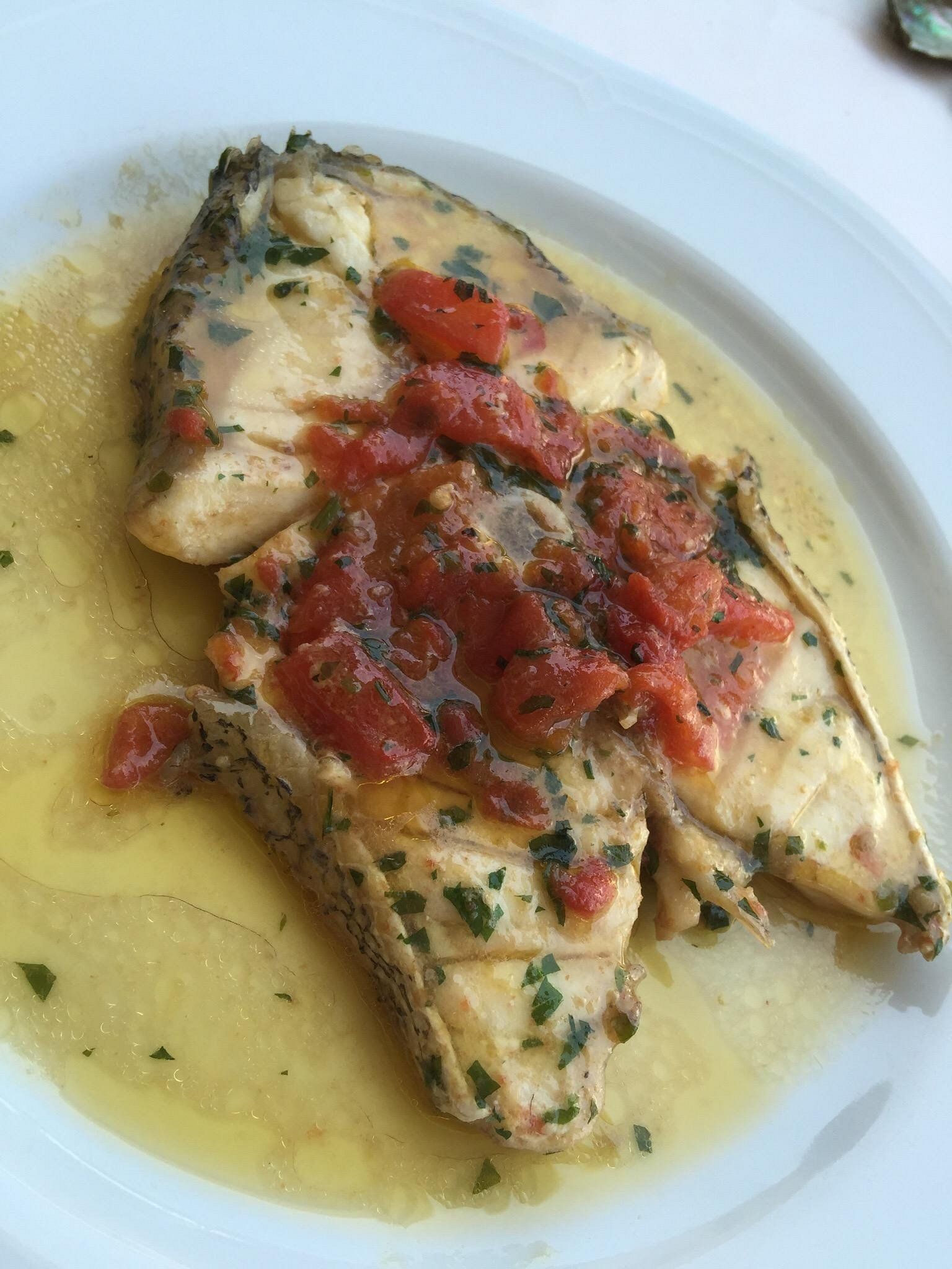 Pesce bianco in salsa cremosa, guarnito con pomodori a pezzi ed erbe aromatiche, servito su un piatto bianco.