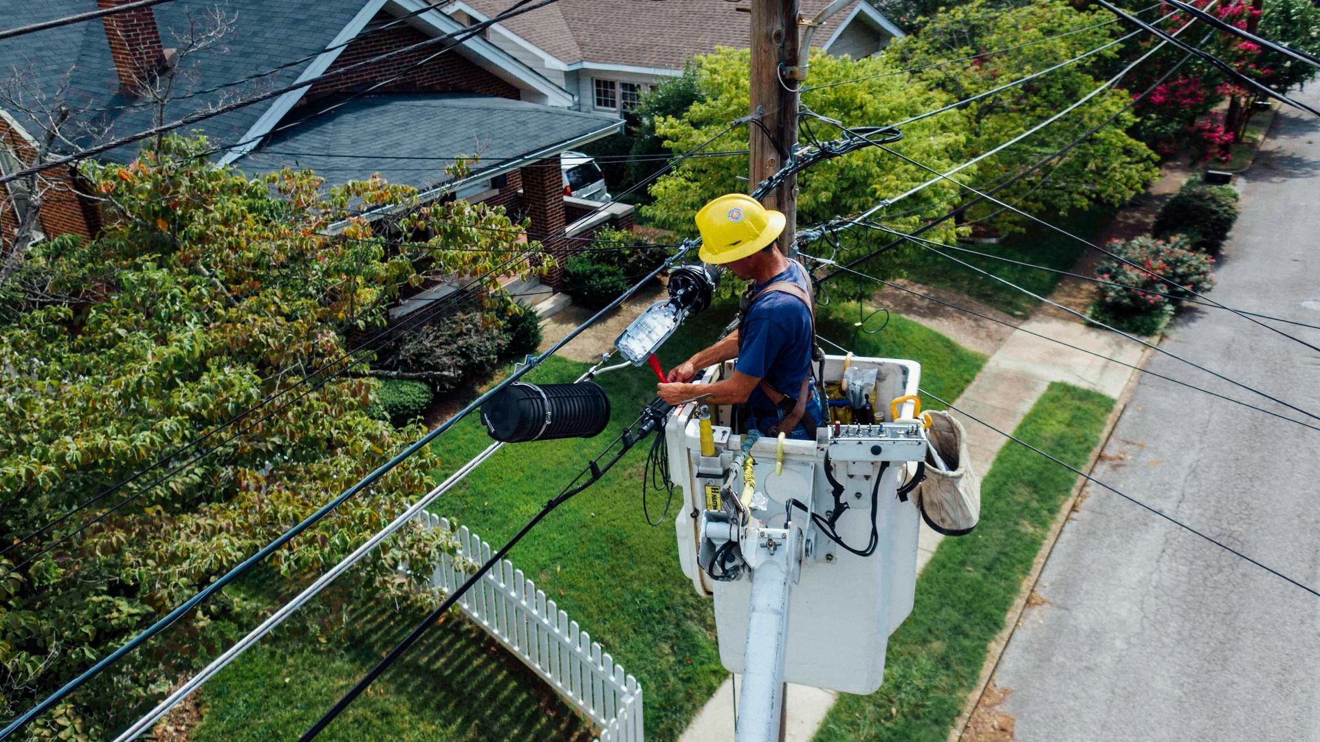A utility worker in a hard hat works on overhead power lines from the bucket of a lift truck in a residential street.