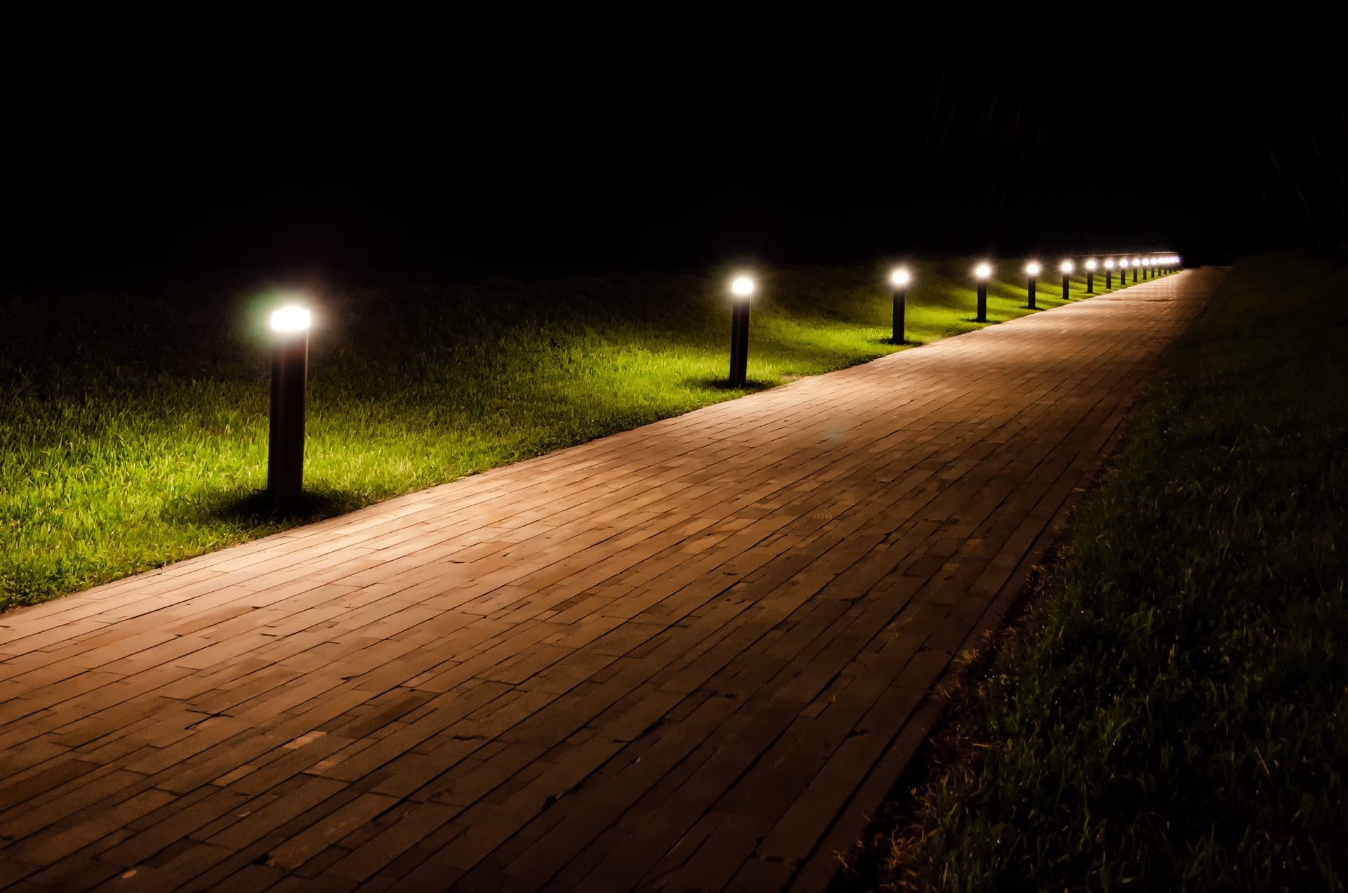 A lit brick path at night with glowing lamp posts along a grassy area