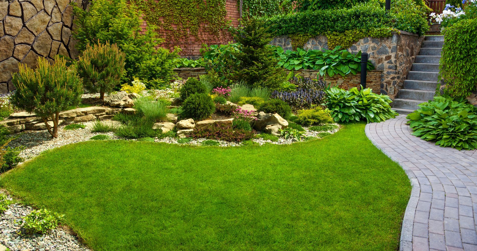 Landscaped garden with green lawn, stone path, flower beds, and steps beside a stone wall