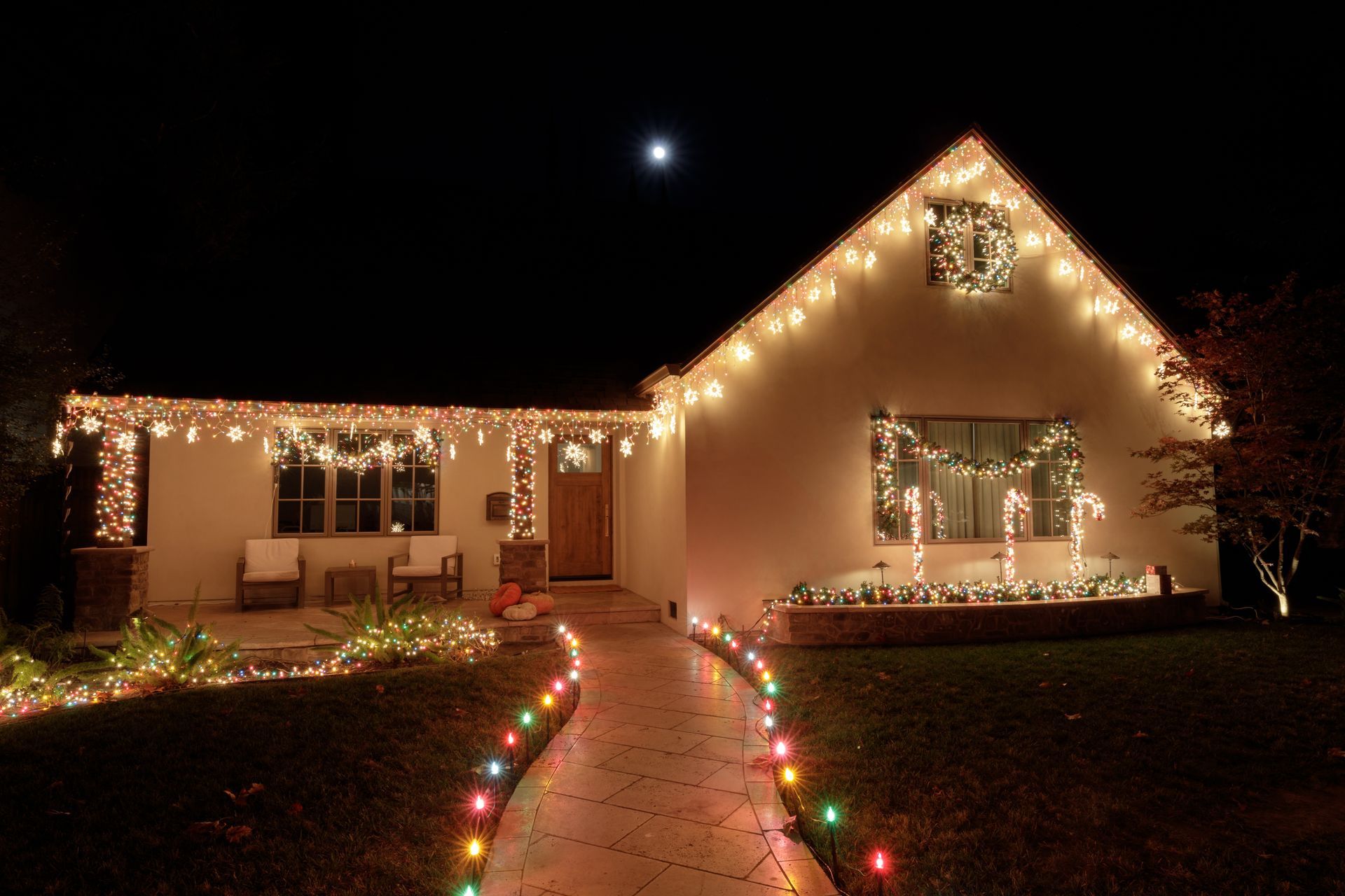 House outlined in warm white Christmas lights, with a lit walkway and a moonlit night sky.