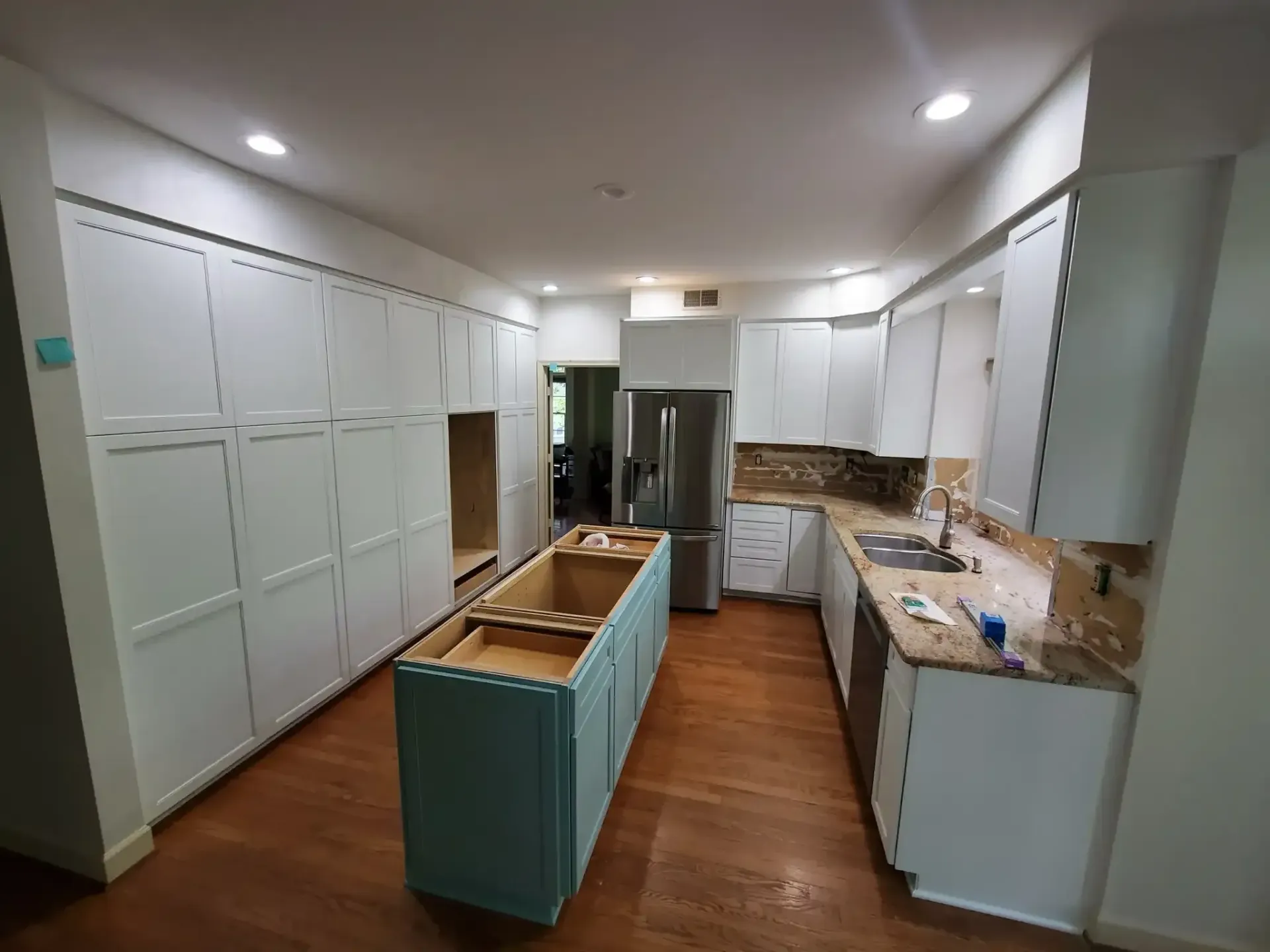 Long white galley kitchen with white cabinets, wood floors, and a light blue island under renovation