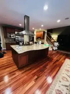 Modern kitchen with dark wood floors, large island, stainless range hood, and staircase in the background