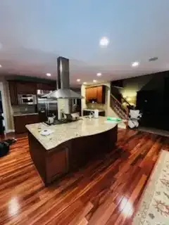 Modern kitchen with island, stainless range hood, dark wood floors, and open staircase nearby