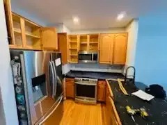 Small kitchen with stainless fridge, wood cabinets, stove, sink, and countertop clutter.