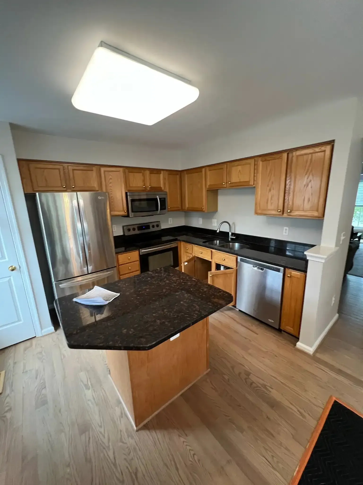 Bright kitchen with wood cabinets, stainless steel appliances, black countertops, and an island under a ceiling light