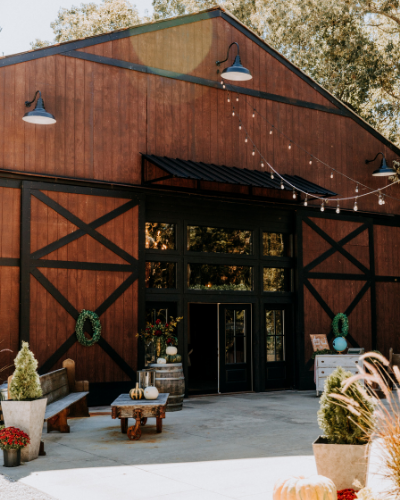 Rustic barn-style venue with wood siding, black trim, string lights, and potted plants at the entrance