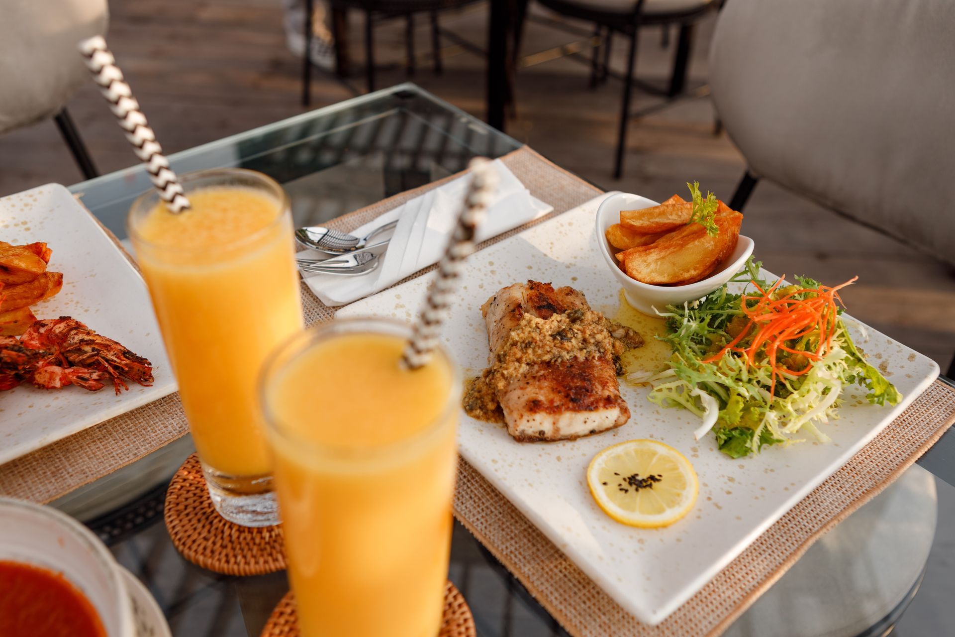A restaurant table setting featuring two tall glasses of vibrant orange juice or smoothie in the blurred foreground, one with a patterned straw. In the background, a square white plate holds a seared white fish fillet with a herb topping, served alongside a lemon wheel, a small side salad, and a bowl of golden potato wedges.