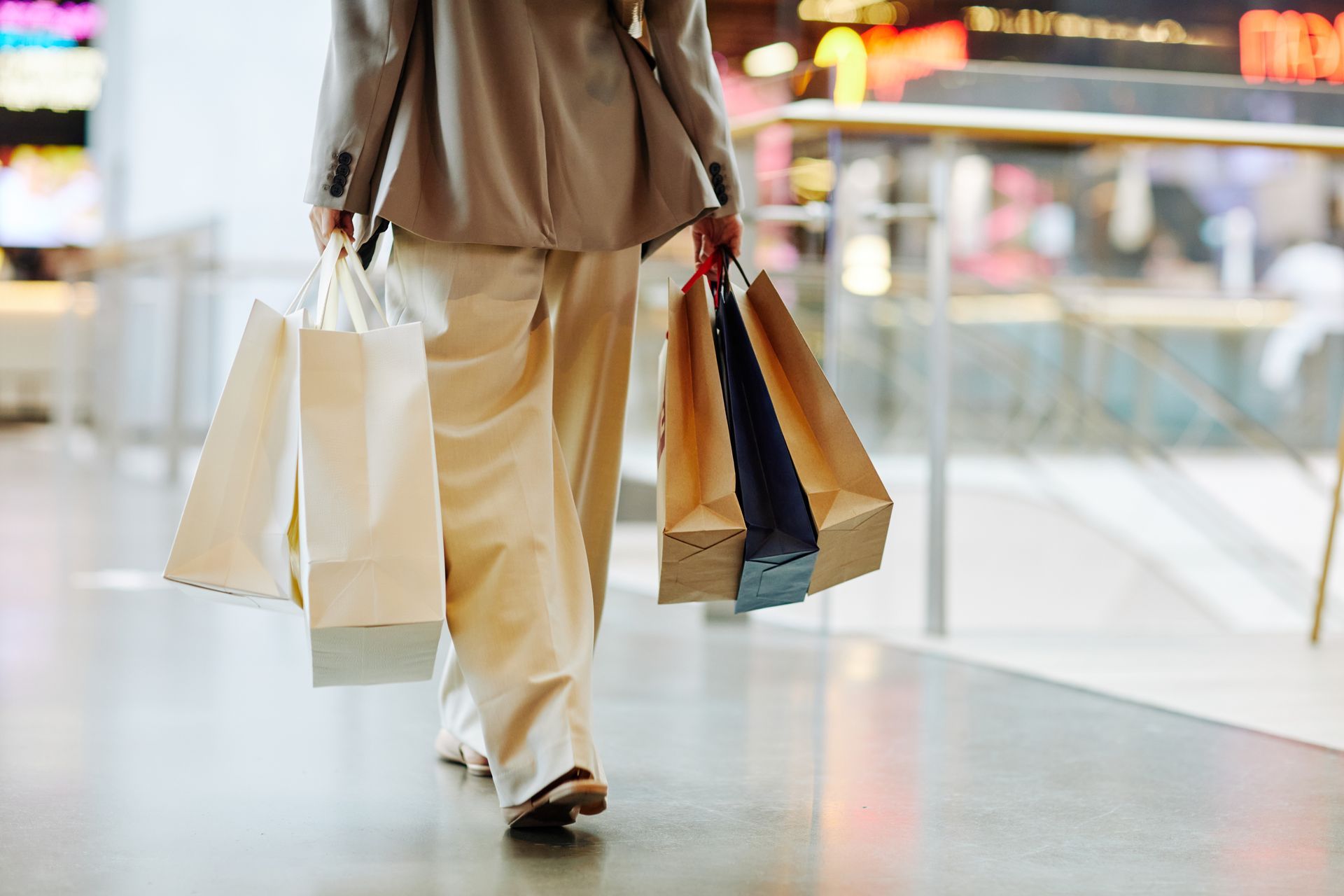 A view from behind of a person walking through a brightly lit shopping mall, carrying multiple white and brown paper shopping bags in both hands. The person is wearing a beige blazer and light-colored wide-leg trousers.