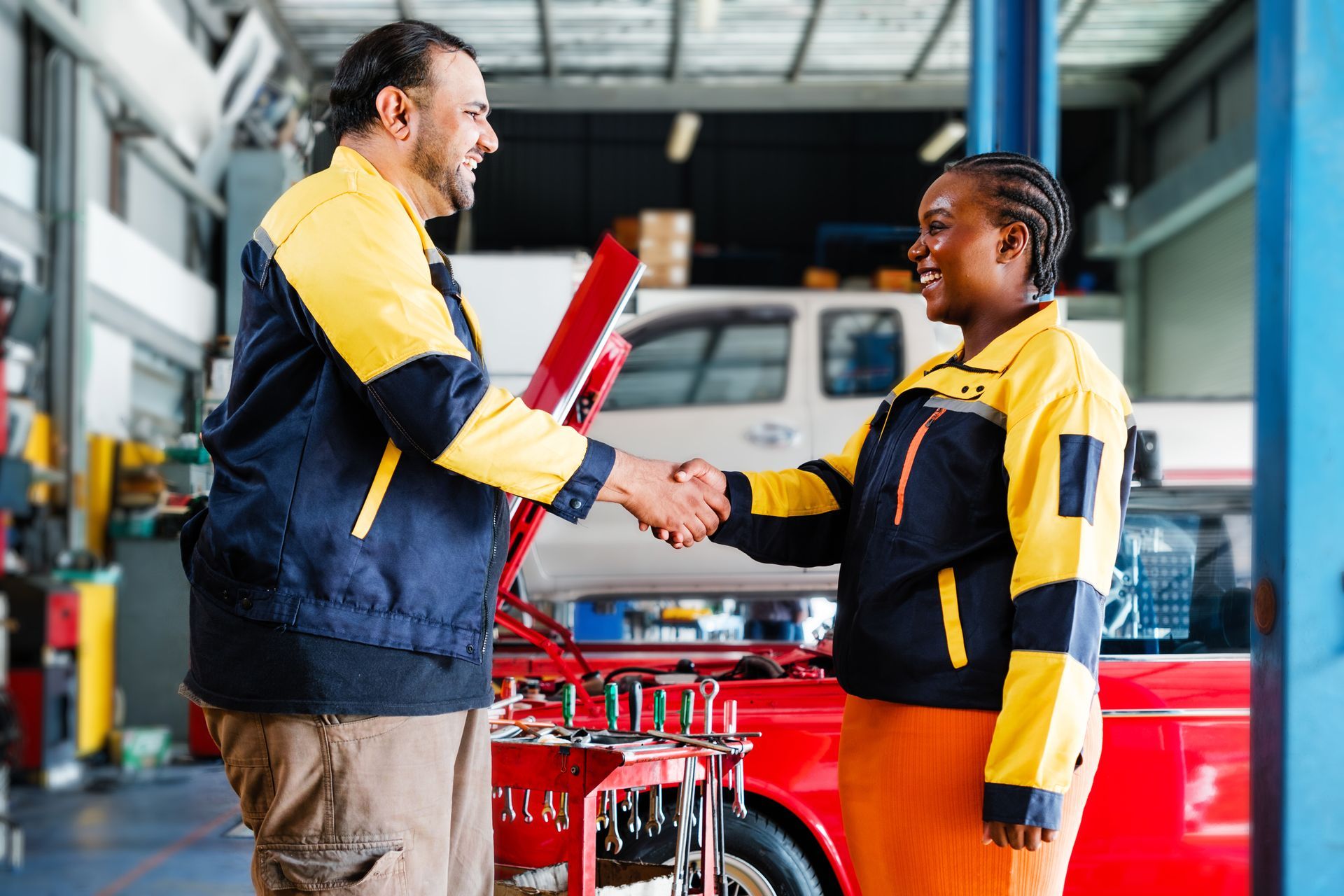 Two mechanics shaking hands in a car repair shop. Both wear navy and yellow uniforms. Two mechanics shaking hands in a car repair shop. Both wear navy and yellow uniforms.