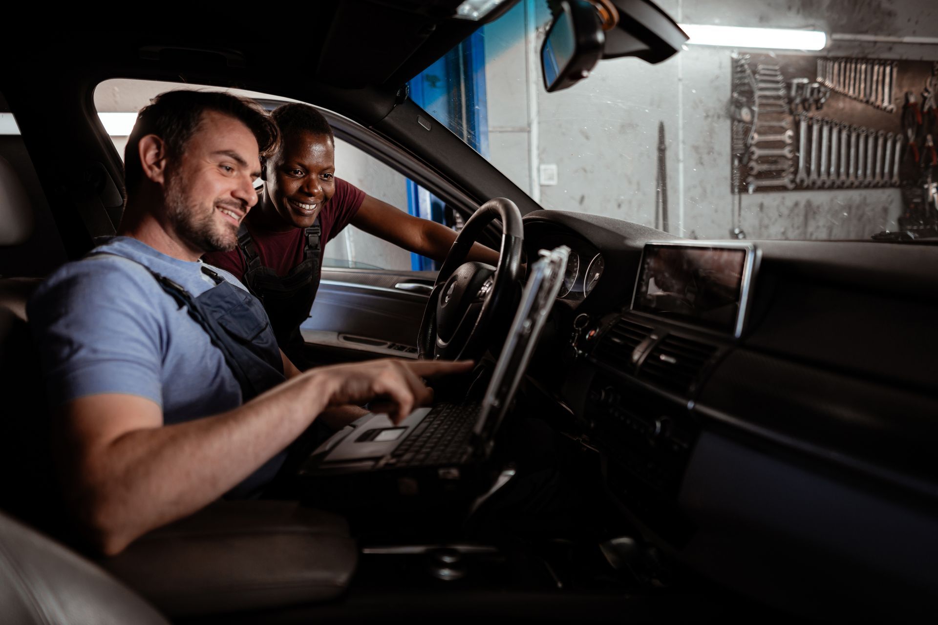 Two mechanics in a car; one points at a laptop on the dashboard, smiling; other mechanic smiles too. Garage setting. Two mechanics in a car; one points at a laptop on the dashboard, smiling; other mechanic smiles too. Garage setting.