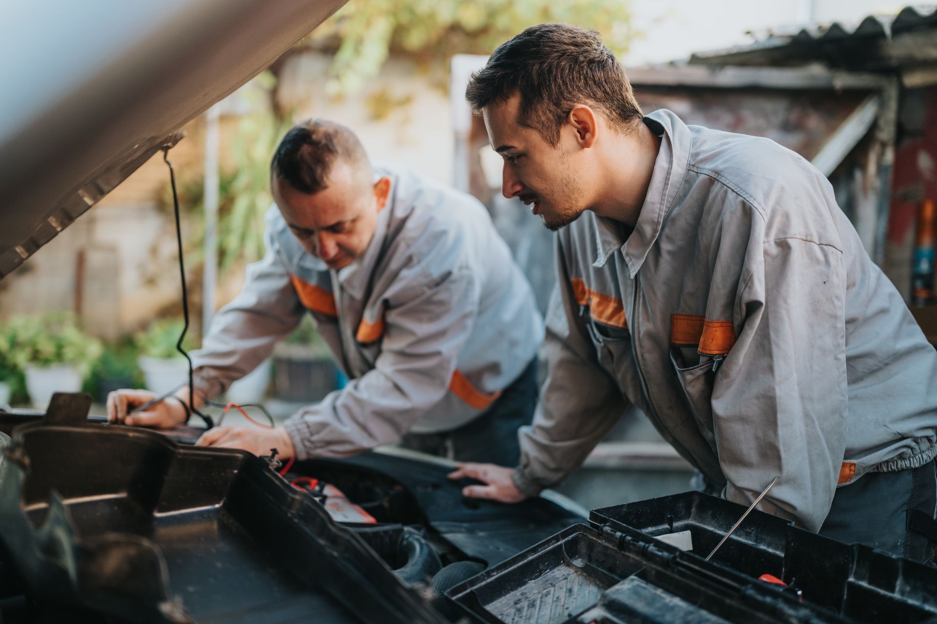 Two mechanics in gray uniforms inspect a car engine with the hood open, outdoors. Two mechanics in gray uniforms inspect a car engine with the hood open, outdoors.