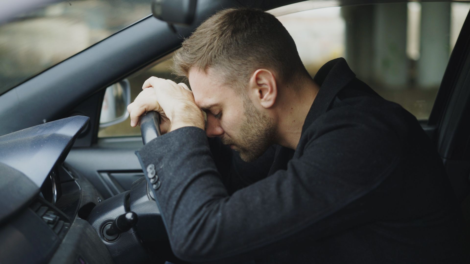 Man resting head on steering wheel inside a car, looking distressed.