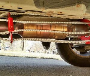 Catalytic converter secured under a car with steel cables and red brackets.