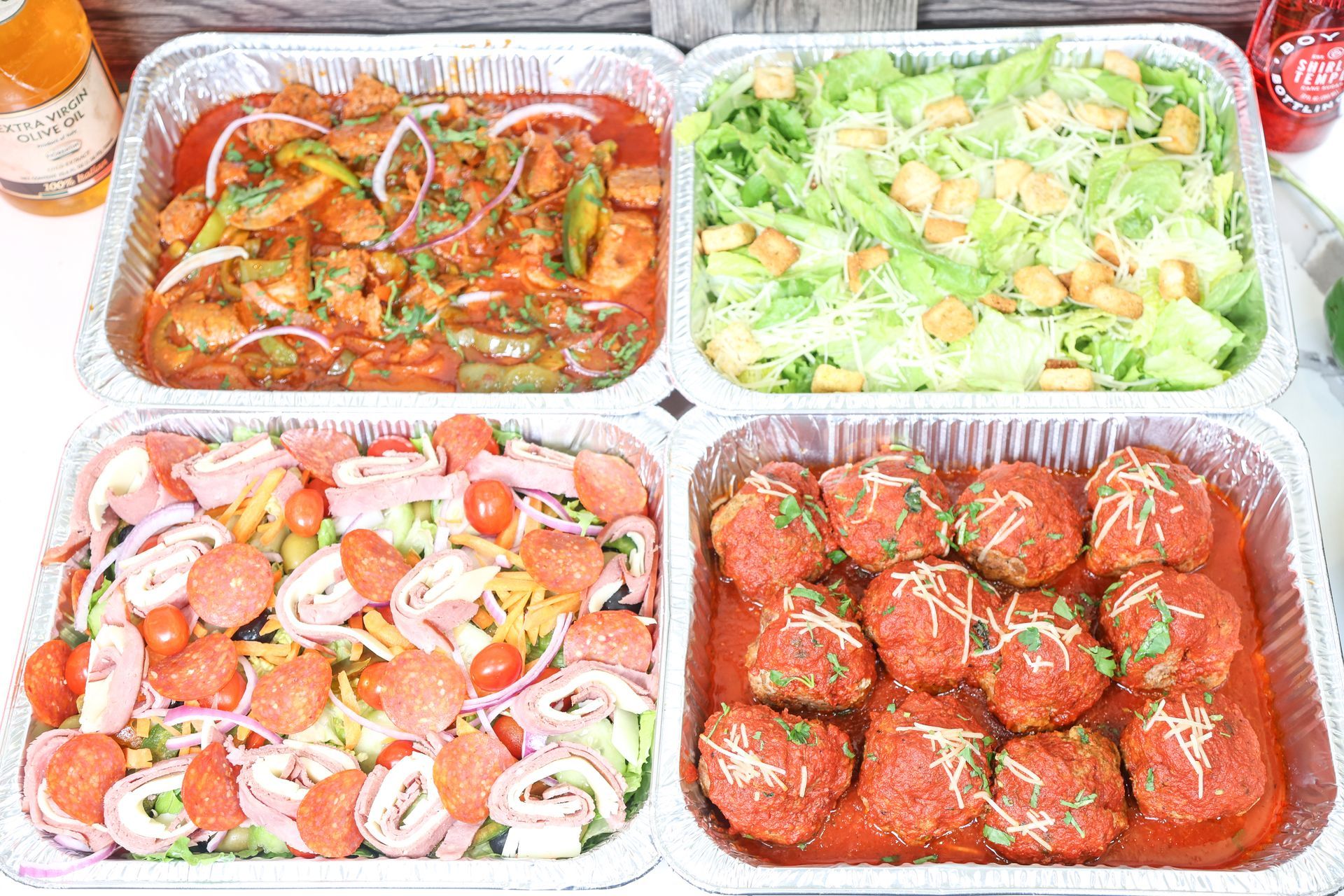 Four foil trays of assorted prepared foods, including saucy meat, vegetables, and salad.