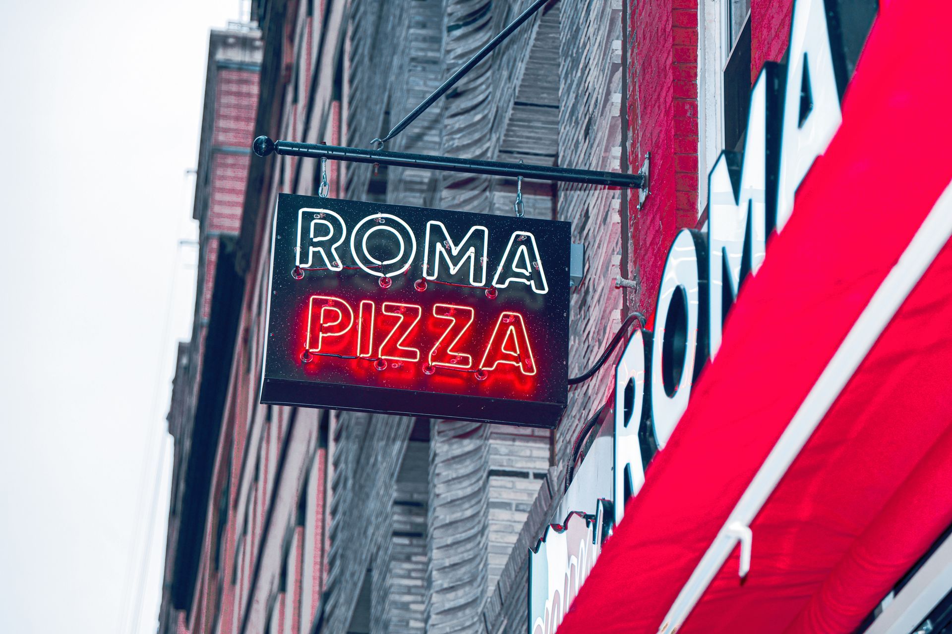 Neon Roma Pizza sign on a city building, with red and white storefront accents