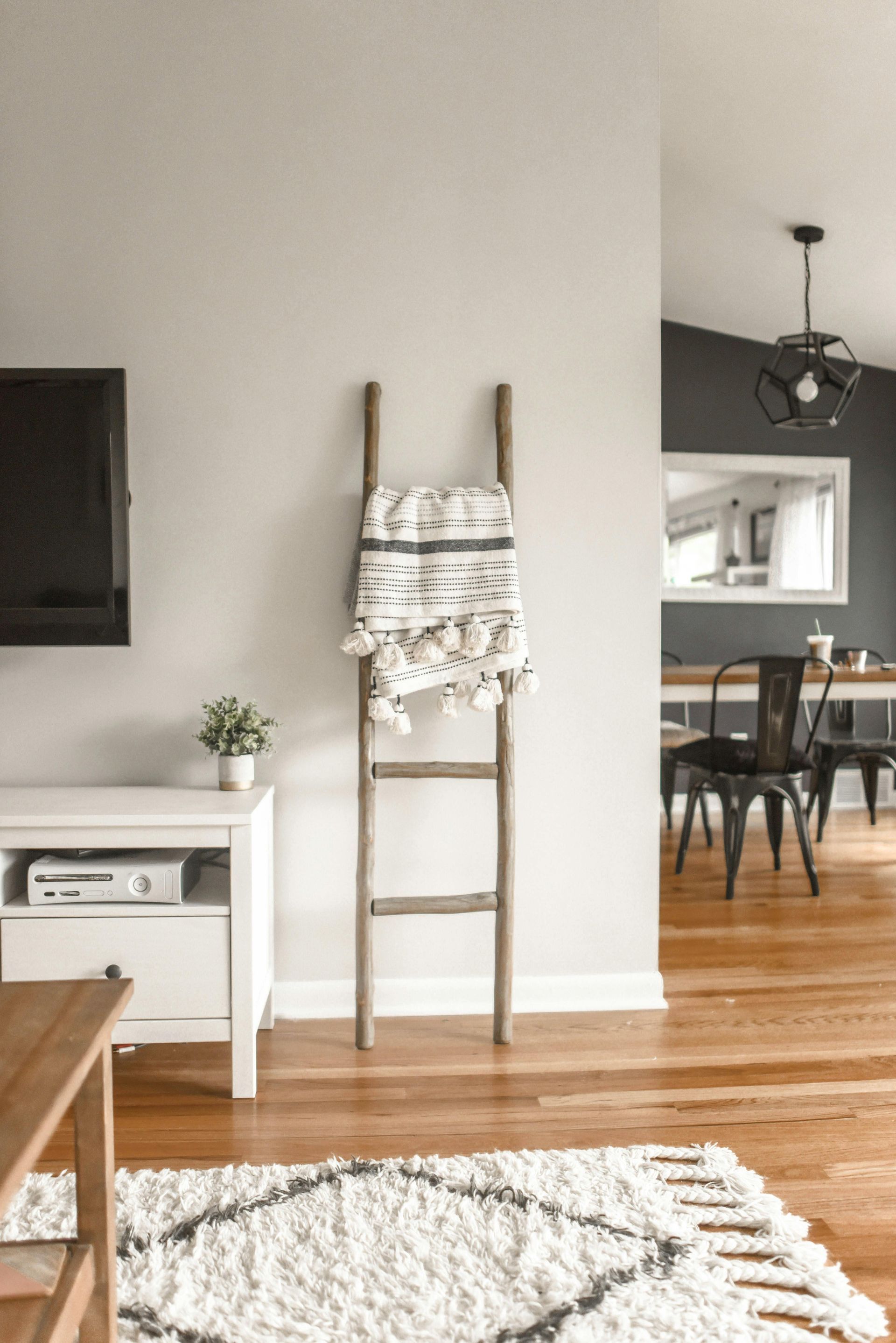 Wooden ladder with decorative blanket against a light gray wall; living room with white rug and dining area visible.