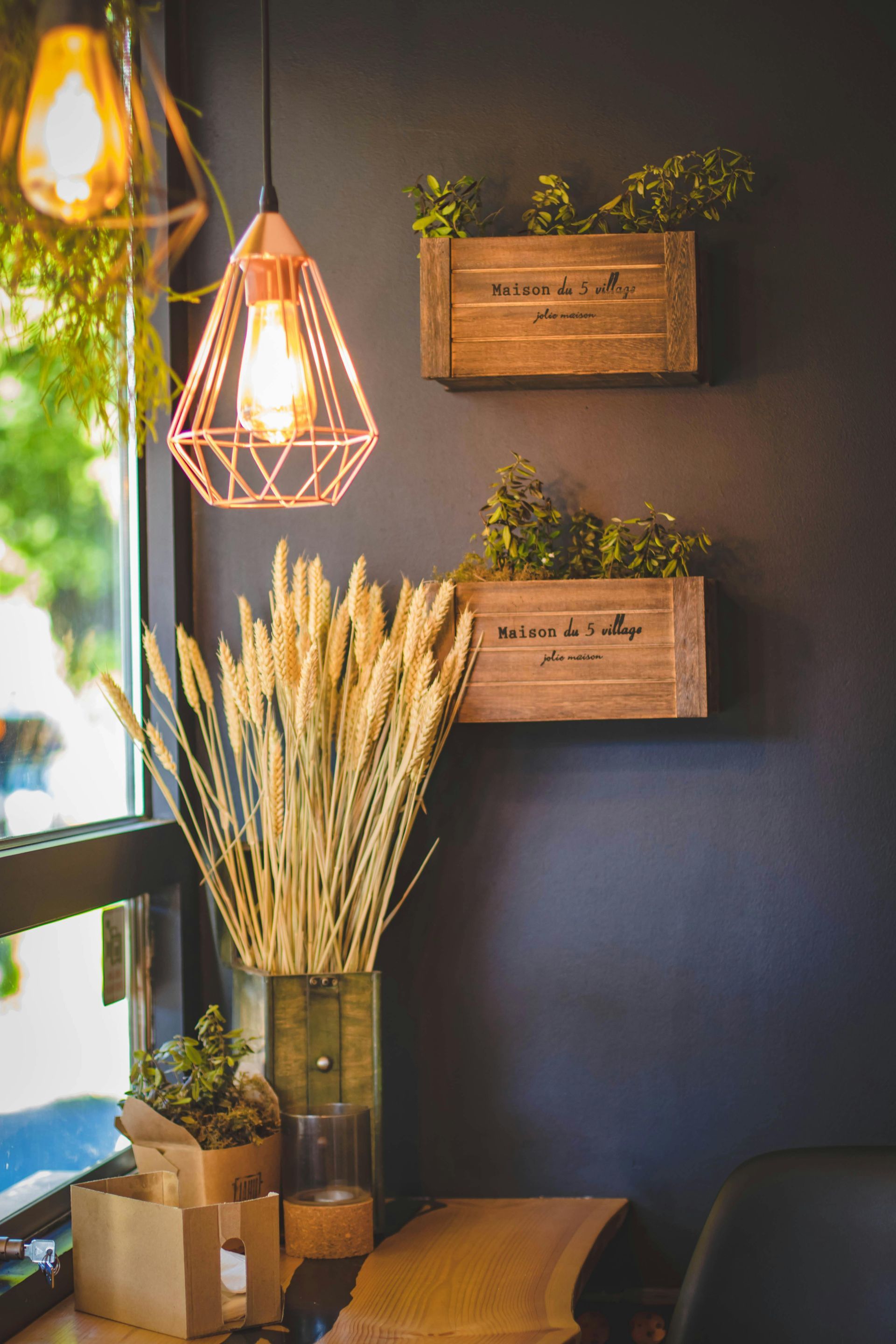 Cafe interior: wheat stalks, wall-mounted wooden planters with greenery, copper geometric pendant lights, and window.