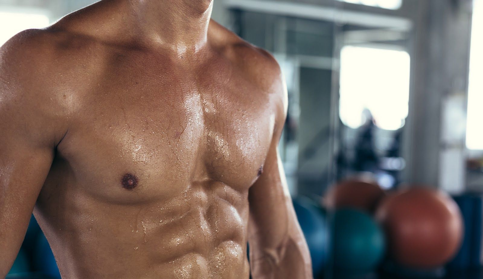 Shirtless athlete torso glistening with sweat in a gym, with exercise balls blurred in the background