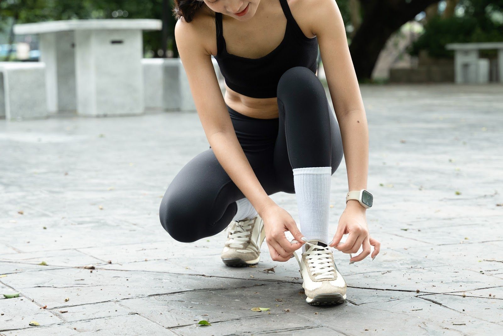 Woman tying shoelaces on white sneakers while kneeling outdoors on a paved path
