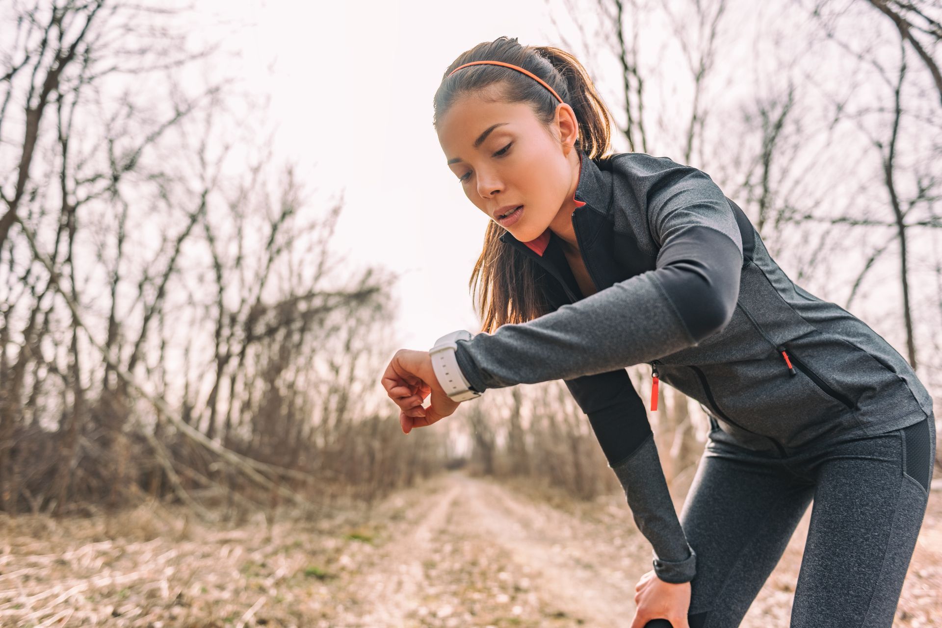 A runner in a dark long-sleeve athletic jacket checks a fitness watch on their wrist while taking a break on a dirt path.