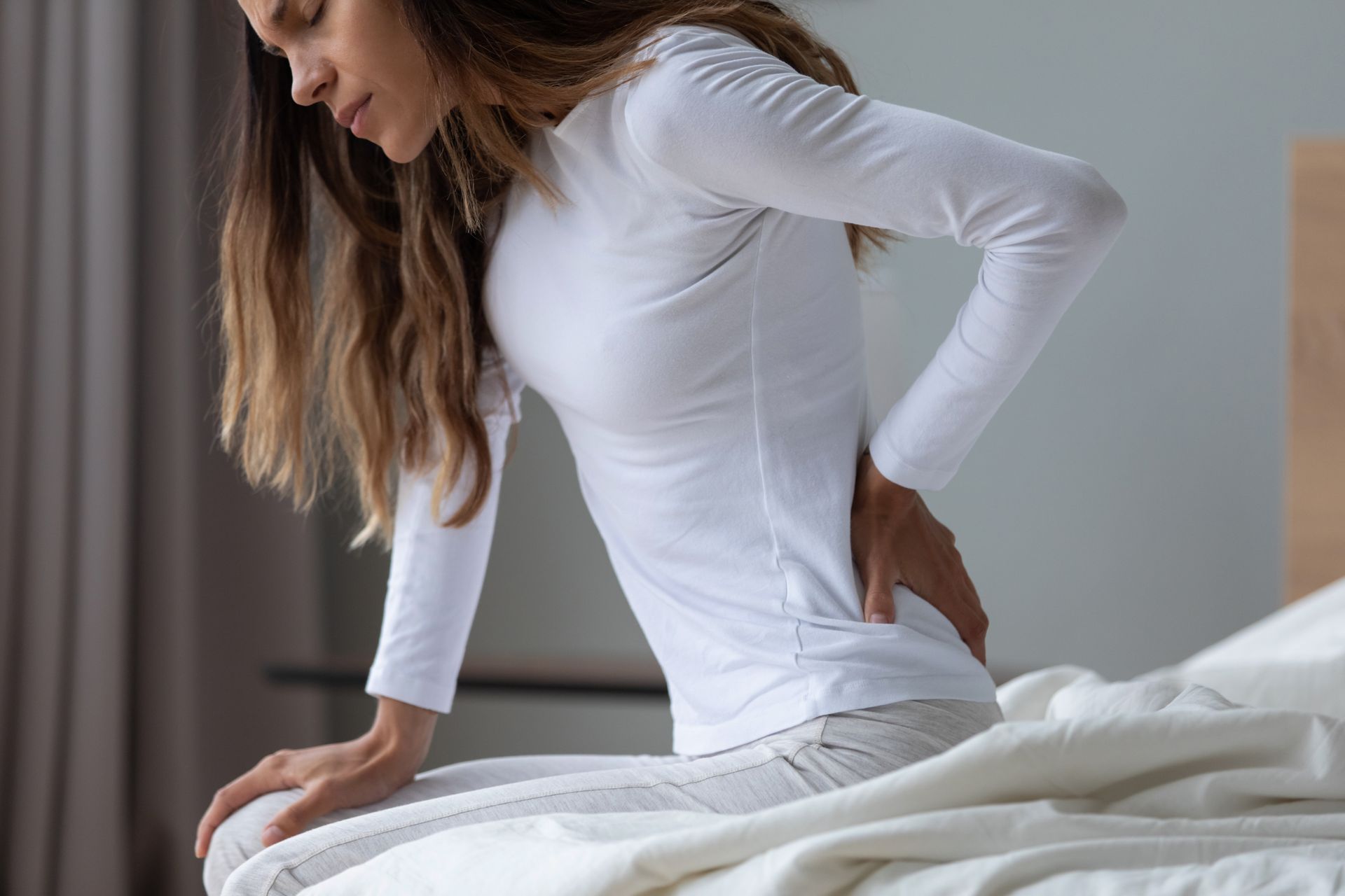 A person sitting on the edge of a bed, holding their lower back with a pained expression.