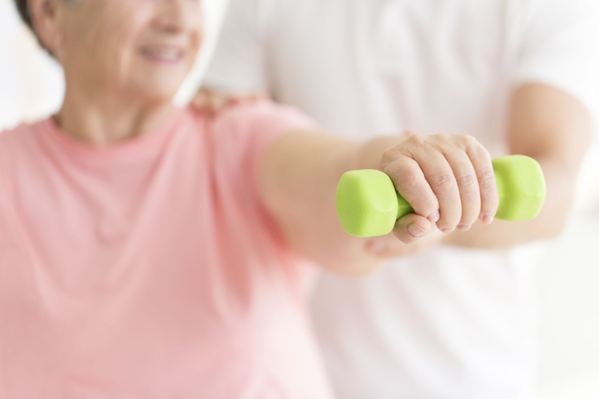 A physical therapist supports a patient as they perform an arm exercise using a small green dumbbell.