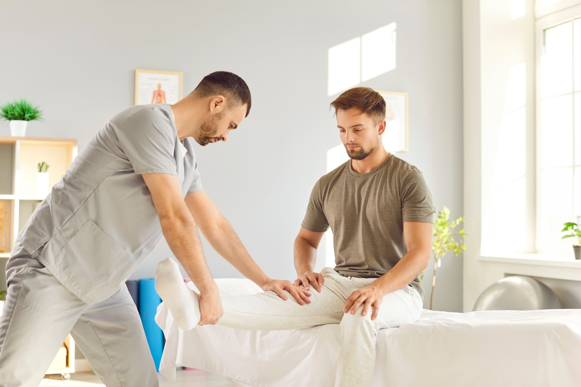 A physical therapist in gray scrubs performs a leg stretch on a seated patient in a bright, modern clinic.