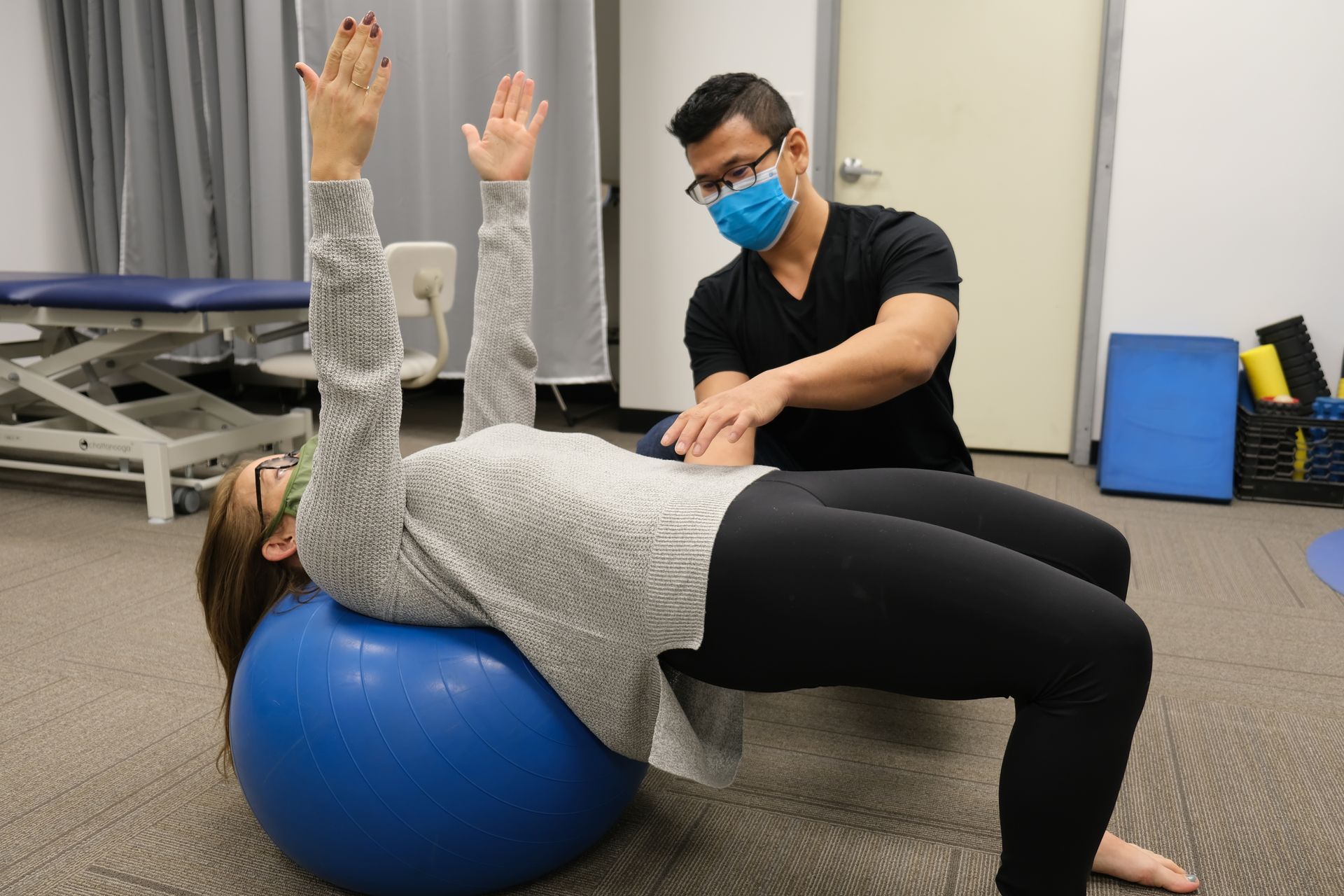 A man is helping a woman do exercises on a blue exercise ball.