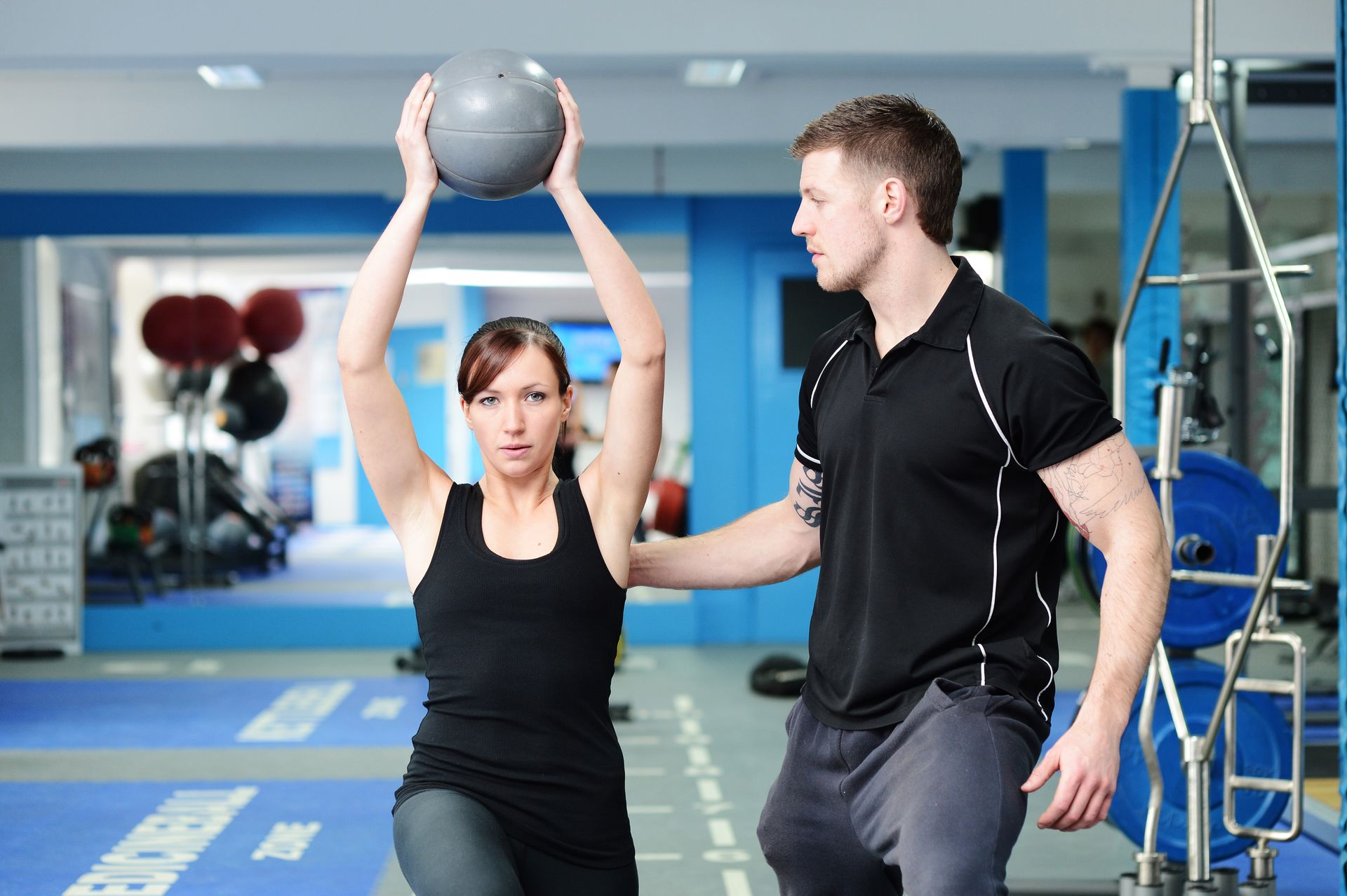 A man is helping a woman with a medicine ball in a gym.