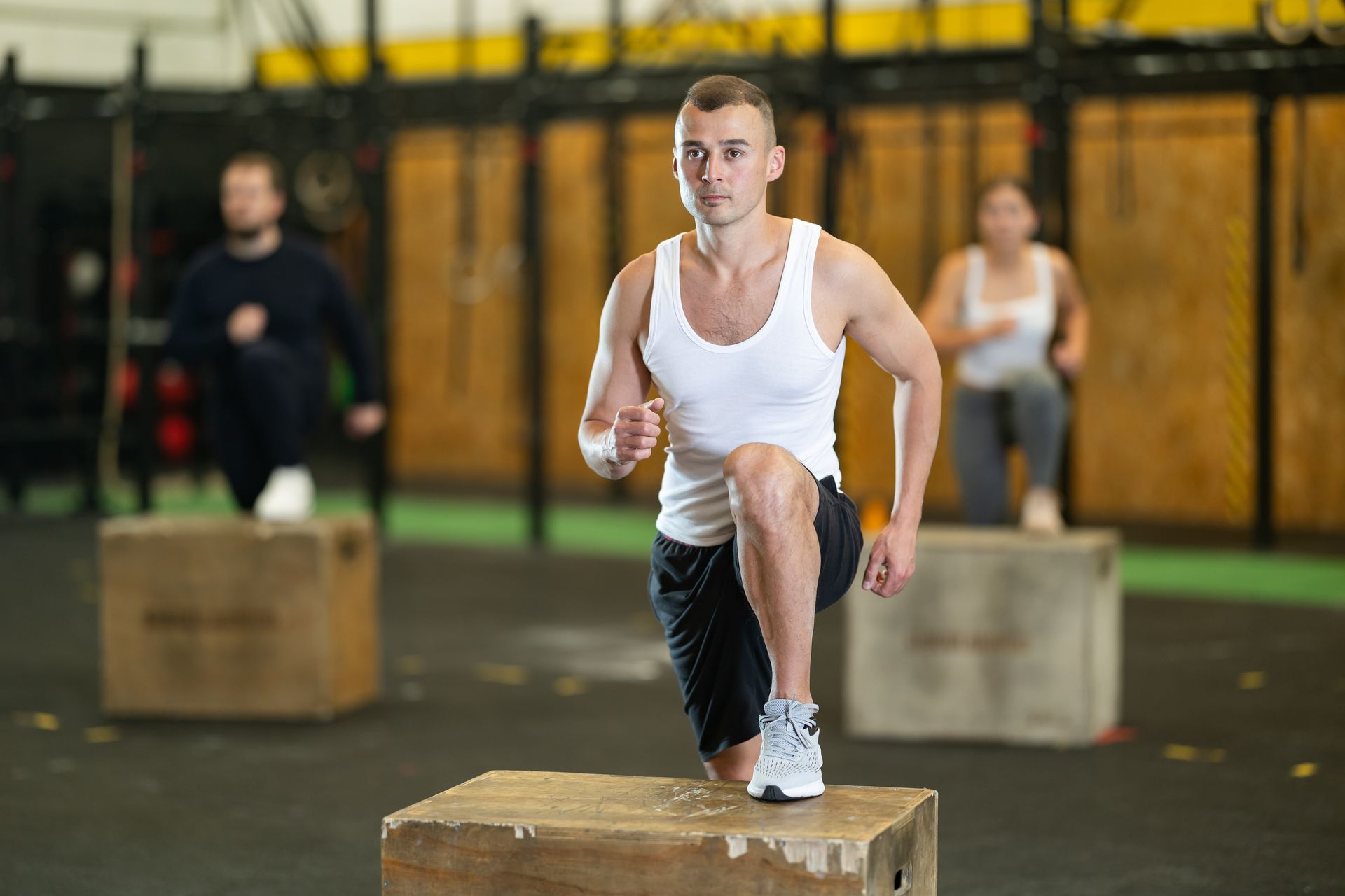 Man in white tank top stepping onto a wooden box at gym; two others exercise in background.