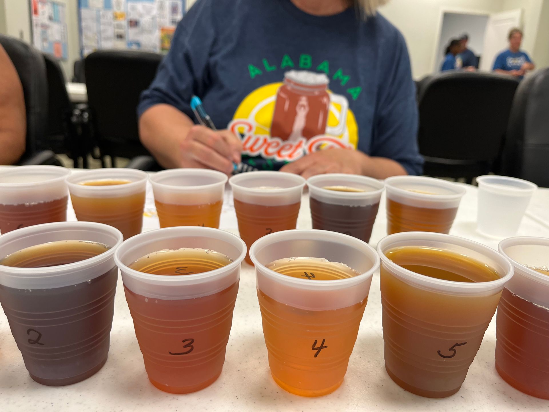 A woman in an alabama shirt sits at a table with plastic cups of different colored liquids