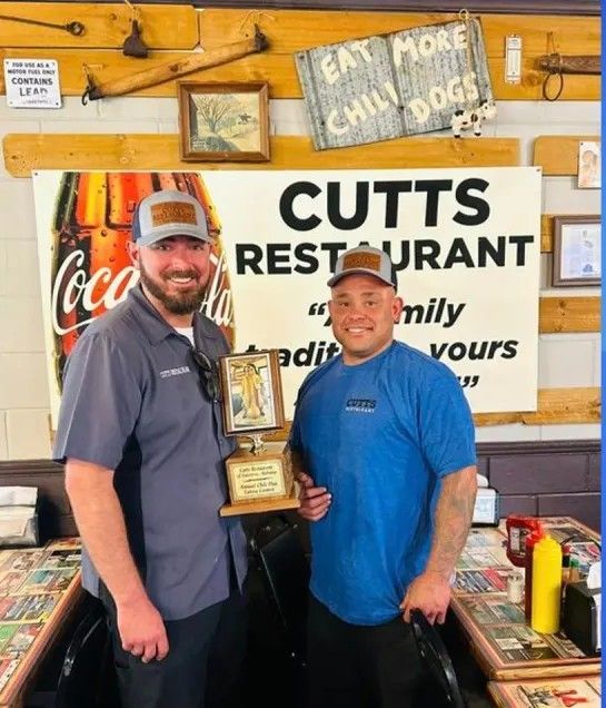 Two men stand inside Cutts Restaurant, holding a trophy. The man on the right is smiling; the restaurant has a Coca-Cola sign.