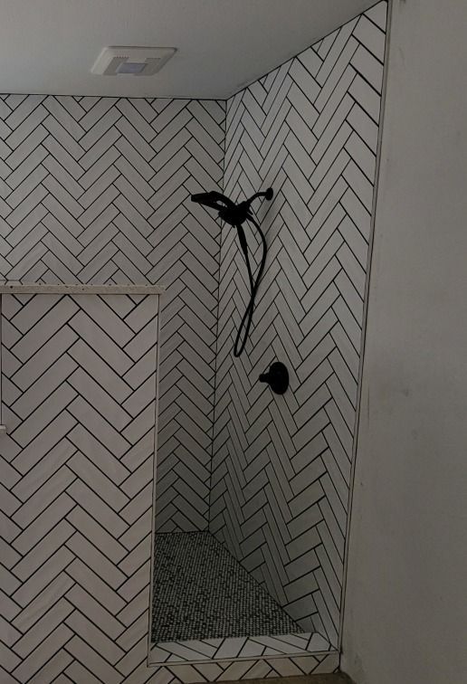 Bathroom shower wall with herringbone tile, exposed shower fittings, and unfinished floor opening