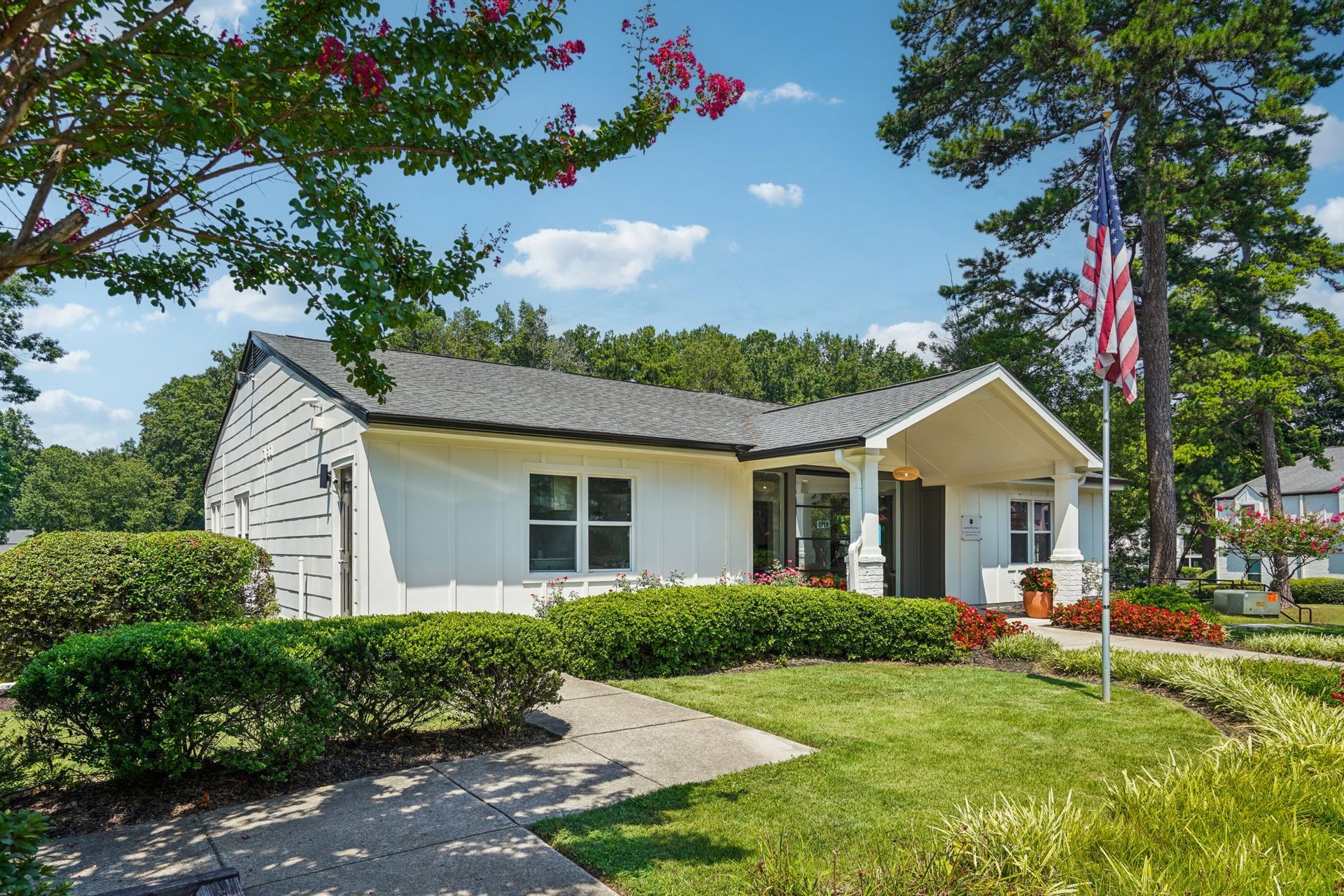 White building with a dark gray roof, shrubs, and a walkway. An American flag flies nearby under a blue sky.