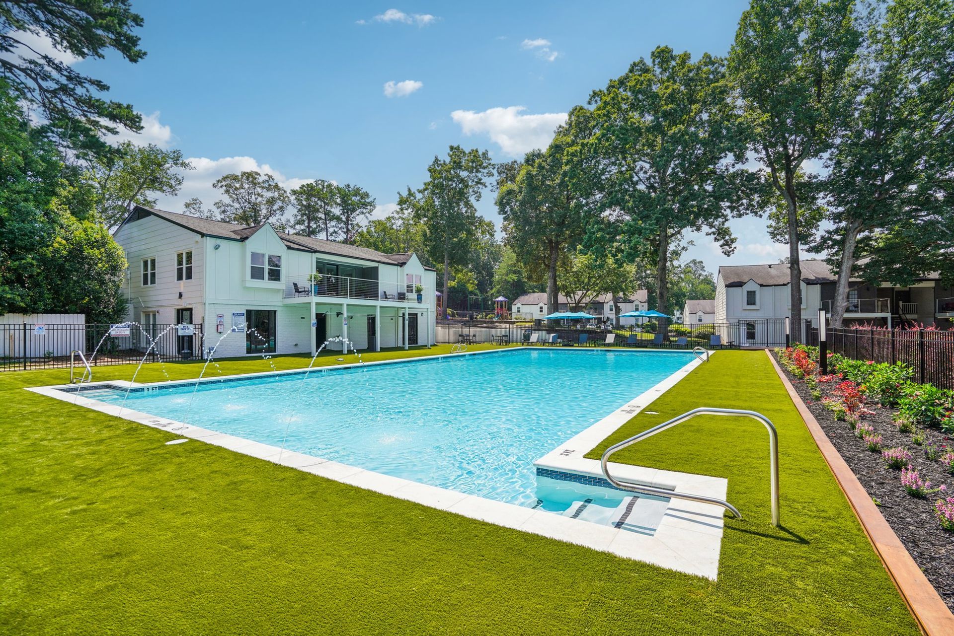 Large rectangular swimming pool with blue water, surrounded by green grass and white buildings under a blue sky.