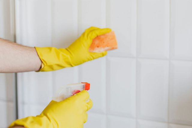 Hands in yellow gloves cleaning white tiles with a sponge and spray bottle