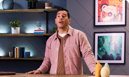 Man in pink shirt at a table in front of shelving with decorative items and framed art.