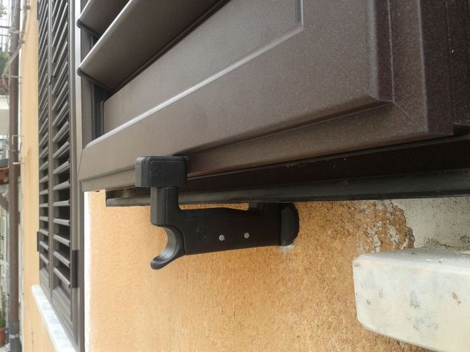 Un gancio di bloccaggio nero per persiana, montato su una parete gialla con superficie ruvida, sotto una persiana marrone.