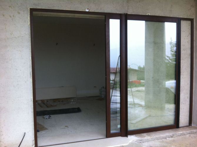 Una porta scorrevole in vetro con cornice marrone è parzialmente aperta, rivelando una stanza non rifinita con pavimento in cemento e pareti spoglie.