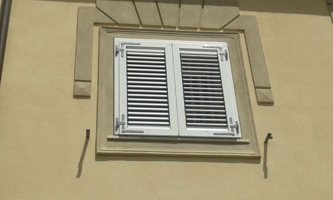 Una finestra con persiane bianche su una parete esterna di colore chiaro, con una modanatura decorativa in pietra sopra il telaio.