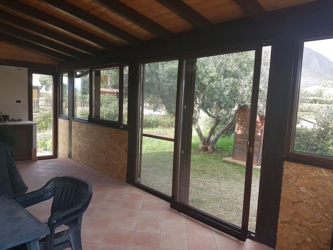 Un patio coperto con soffitto a travi in legno, pavimento piastrellato, sedia in plastica e grandi finestre in vetro che si affacciano su un giardino.