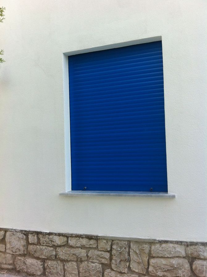 Una serranda avvolgibile di un blu brillante copre una finestra su una parete esterna bianca, sopra una base in pietra.