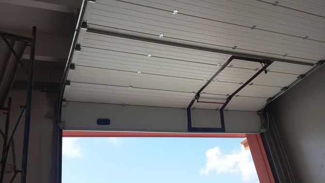 Un portone industriale bianco è parzialmente sollevato, rivelando un cielo azzurro brillante all'esterno.