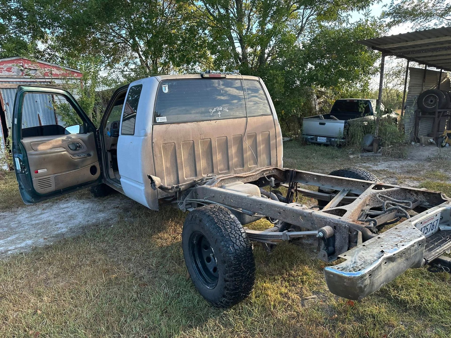 A silver truck cab with the driver door open, parked on grass without a truck bed, revealing the rear frame and axle.