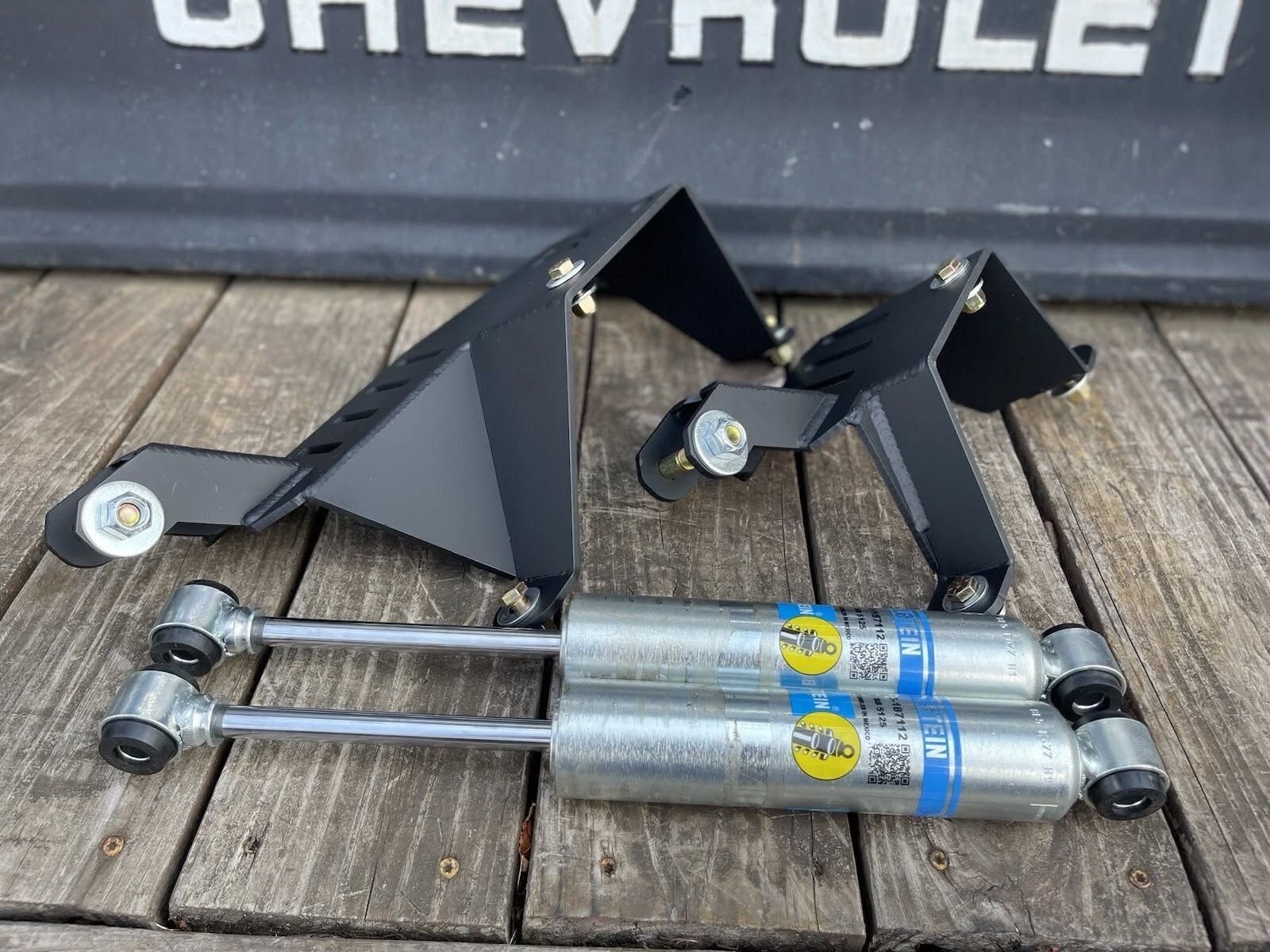 Two metal suspension brackets and two Bilstein shock absorbers arranged on a wooden surface with a Chevrolet logo above.