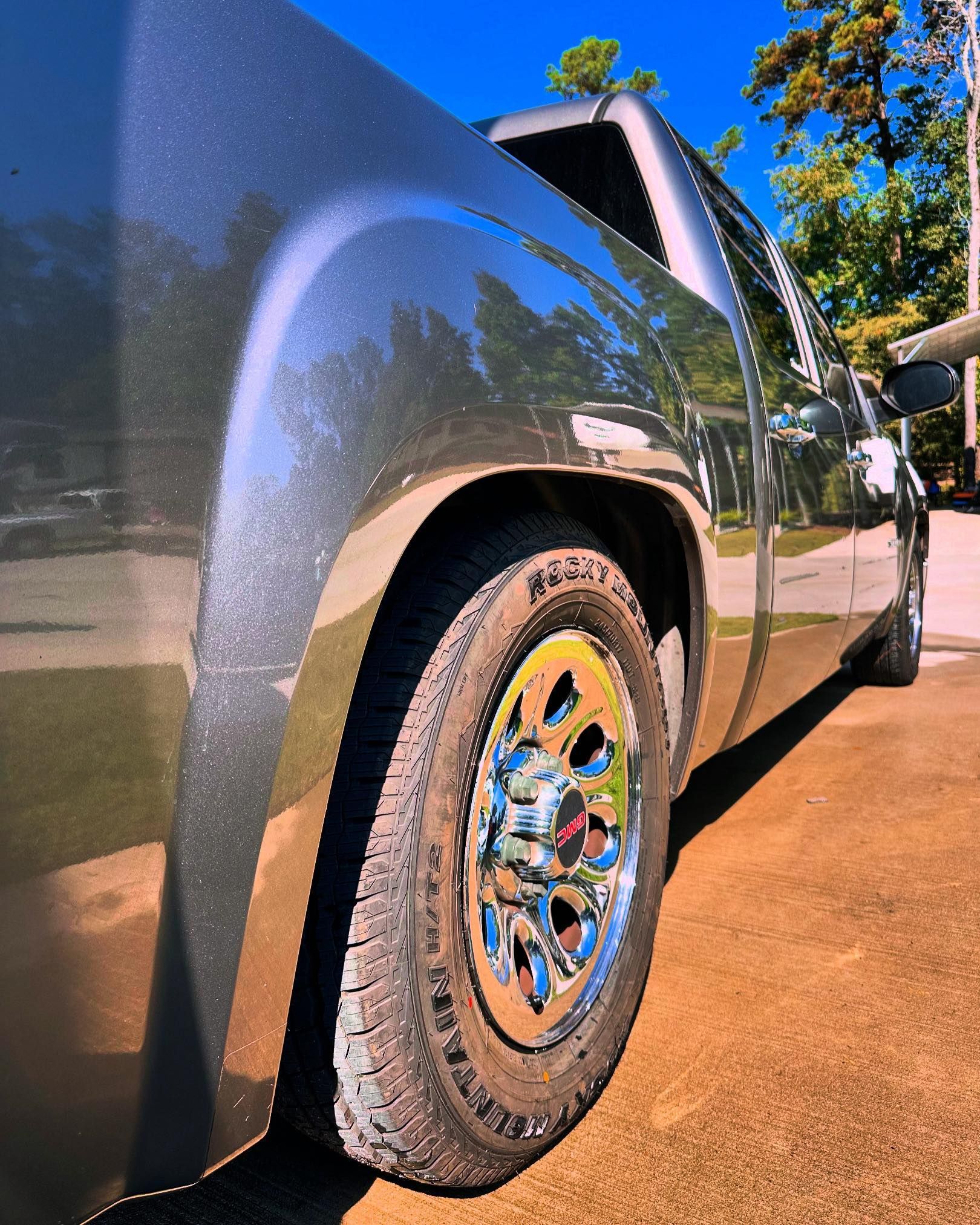 A side view of a dark grey pickup truck, highlighting a chrome wheel and tire parked on a paved driveway in sunlight.