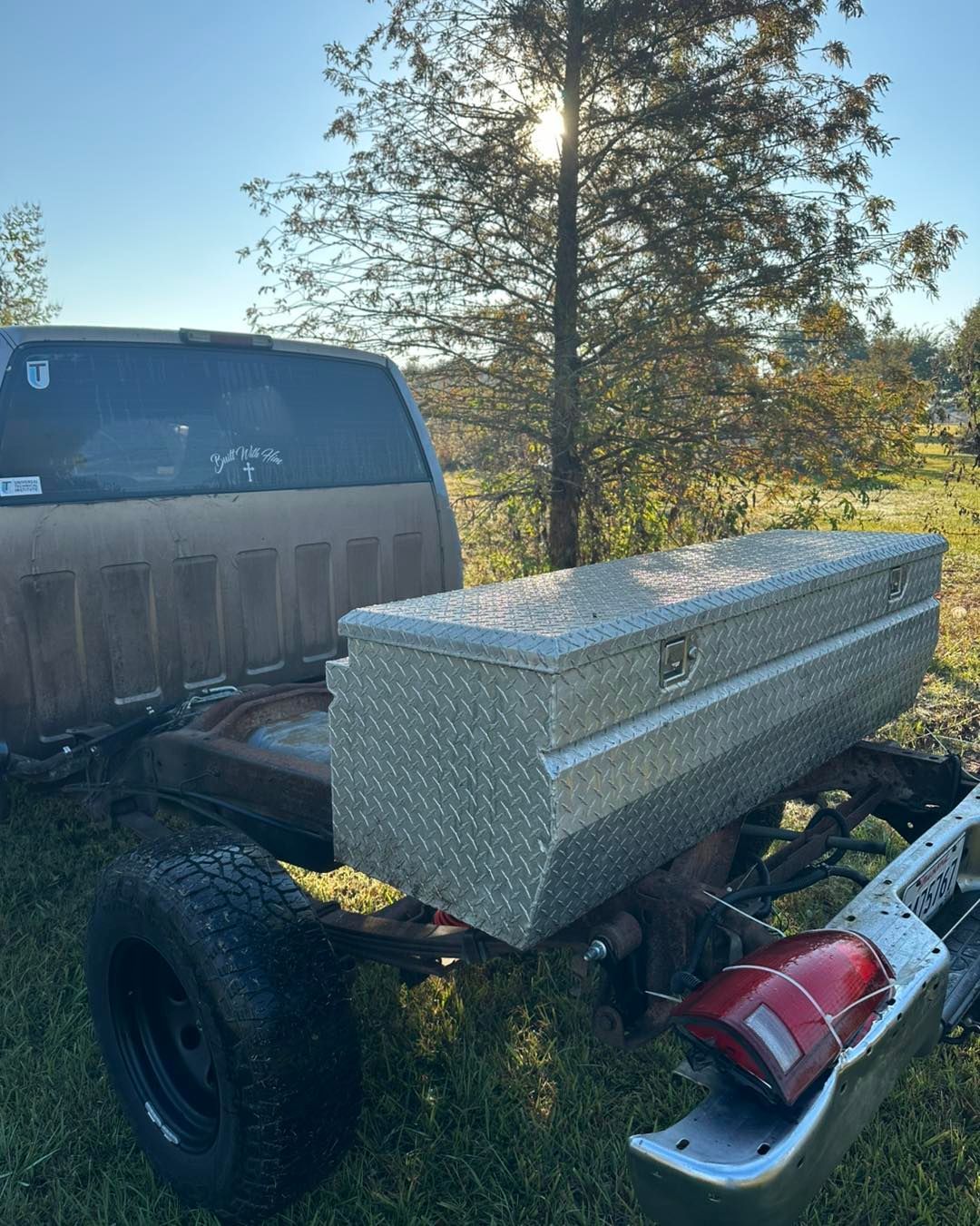 A silver diamond-plate toolbox sits on the bare frame of a truck without a bed, parked in a grassy field.